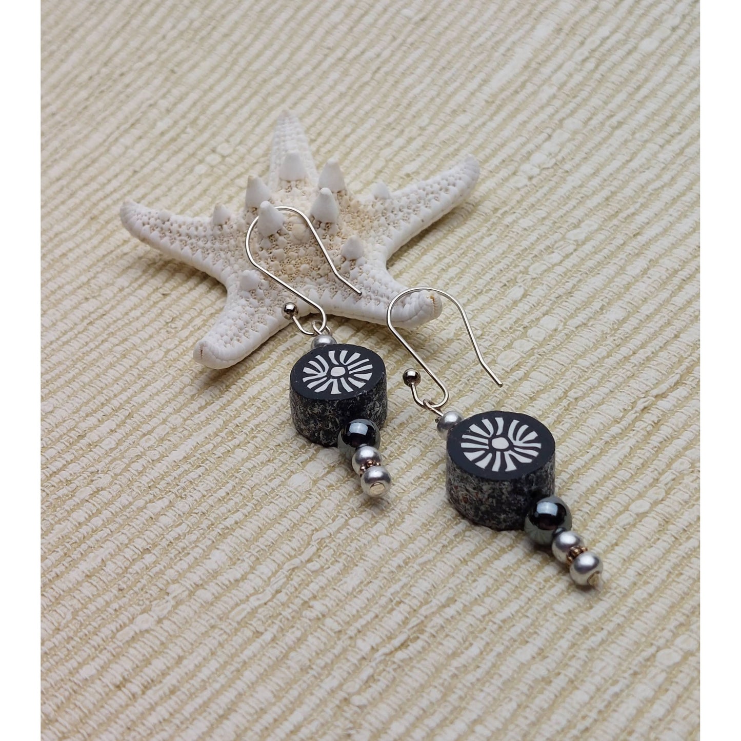 Polymer clay sunburst disk and hematite gemstone drop earrings with sterling silver ear wires leaning on starfish on linen background