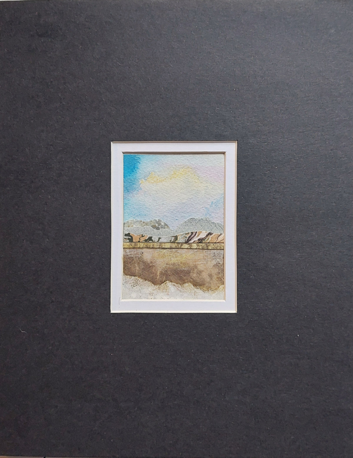 A small framed watercolor landscape collage matted in a black and white double mat. The collage depicts a scenic view with blue sky, distant mountains, and a foreground of brown and beige tones.